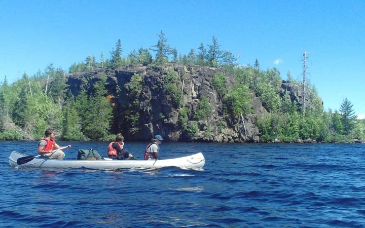 Three people paddle a canoe on blue water past a rocky shore.